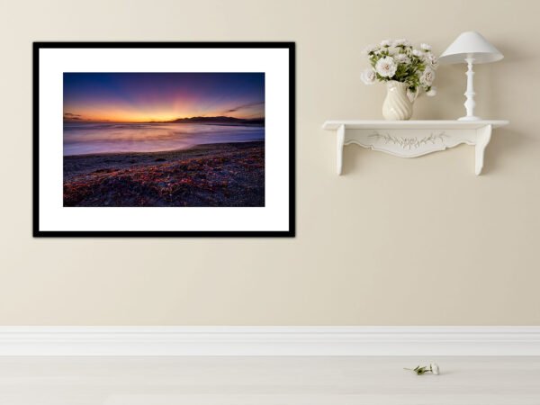 Framed sunset photograph on wall near shelf with vase.