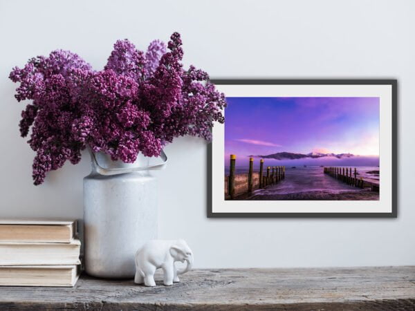 Lilacs in vase with framed scenic photo.